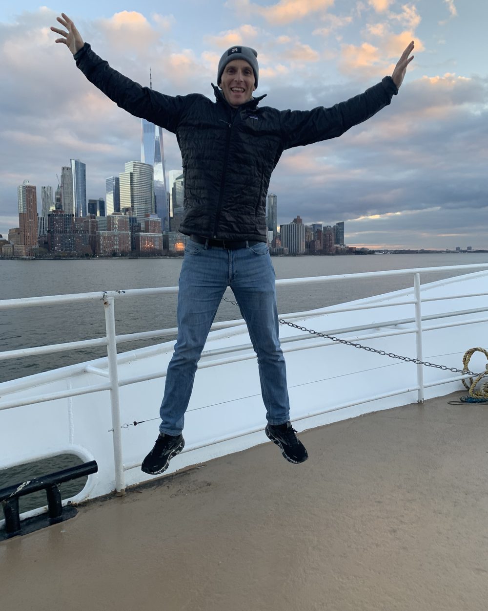 Jay jumping with arms spread on a ferry deck in front of the New York skyline